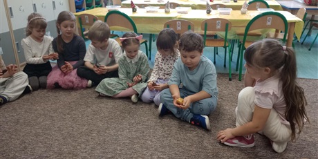 Powiększ grafikę: Dzieci siedzą na dywanie i oglądają bursztyn. Mają w rękach bryłki bursztynu.