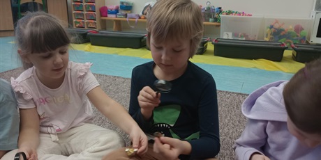 Powiększ grafikę: Grupa dzieci ogląda bursztyn. Chłopiec trzyma w ręku lupę. Wszyscy są radośni.