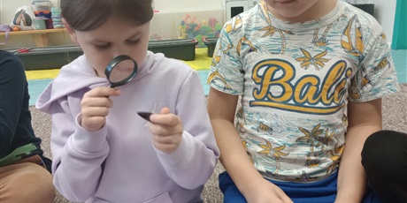 Powiększ grafikę: Grupa dzieci ogląda bursztyn. Dziewczynka trzyma w ręku lupę. Wszyscy są radośni.