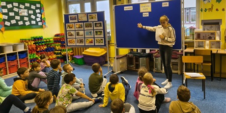 Powiększ grafikę: Grupa dzieci siedzi na dywanie w świetlicy zwrócona w stronę tablicy informacyjnej. Przed tablicą stoi nauczycielka, która  wskazuje ręką na granatową tablicę z napisem Jesienne Dary.