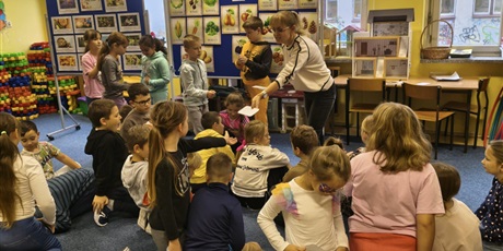 Powiększ grafikę: Nauczycielka rozdaje dzieciom karty do gry w jesienne domino. Część dzieci siedzi na dywanie, dwóch chłopców i trzy dziewczynki stoją. W tle znajdują się dwie granatowe tablice z informacjami o jesien