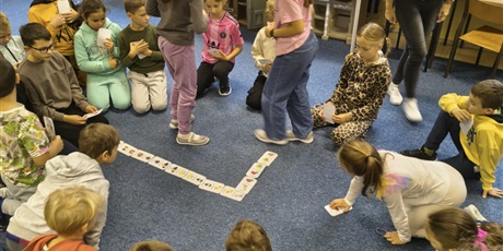 Powiększ grafikę: Grupa dzieci znajduje się na dywanie w świetlicy. W rękach trzymają karty do jesiennego domina. Kolejno rozkładają karty na dywanie.