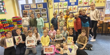 Powiększ grafikę: Grupa dzieci wraz z nauczycielką pozują do zdjęcia. Niektóre dzieci trzymają w rękach ilustracje charakteryzujące jesie, np. słoneczniki, wrzost, przetwory. W tle znajdują się dwie granatowe tablice