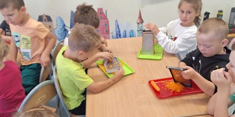 Powiększ grafikę: Dzieci ścierają na tarce marchewkę. Chłopiec trzyma tarkę. Dziewczynka na niego patrzy. W tle widać inne dzieci, które tarkują inne warzywa.