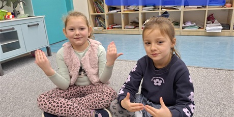 Powiększ grafikę: Dziewczynki siedzą na dywanie. Są uśmiechnięte ale skupione. Pokazują rękami słowa w języku migowym - dzień dobry.