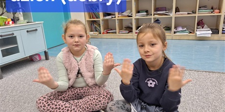 Powiększ grafikę: Dziewczynki siedzą na dywanie. Są uśmiechnięte ale skupione. Pokazują rękami słowa w języku migowym - dzień dobry.