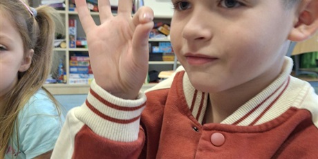 Powiększ grafikę: Chłopiec siedzi na dywanie i uczy się nowego słowa w języku migowym. Pokazuję ręką słowo - dobrze