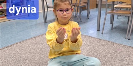 Powiększ grafikę: Dziewczynka siedzi na dywanie i pokazuje pierwszą część wyrazu dynia w języku migowym. Jest bardzo skupiona.