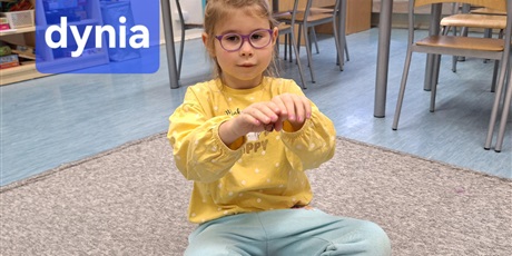 Powiększ grafikę: Dziewczynka siedzi na dywanie i pokazuje drugą część wyrazu dynia w języku migowym. Jest bardzo skupiona.