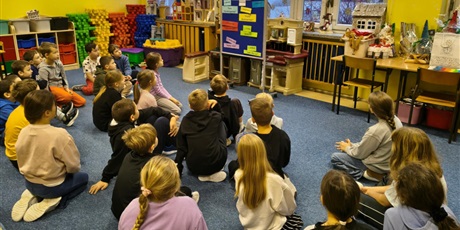 Powiększ grafikę: Grupa dzieci siedzi na dywanie w świetlicy zwrócona w stronę granatowej tablicy z kolorowymi napisami WESOŁYCH ŚWIĄT w różnych językach świata.