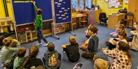 Powiększ grafikę: Chłopiec w zielonej bluzce wiesza karteczki z wyrazami na granatowej tablicy. Reszta dzieci siedzi na dywanie i obserwuje wykonanie zadania przez chłopca.