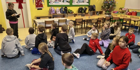 Powiększ grafikę: Dzieci siedzą na dywanie w świetlicy. Jedna dziewczynka stoi i czyta zadanie z kalendarza adwentowego.