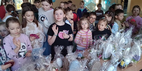 Powiększ grafikę: Grupa dzieci na korytarzu szkolnym podczas kiermaszu wielkanocnego. Dzieci oglądają i kupują ozdoby wielkanocne. Na stole rozłożone są dekoracje.