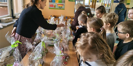 Powiększ grafikę: Grupa dzieci na korytarzu szkolnym podczas kiermaszu wielkanocnego. Dzieci oglądają i kupują ozdoby wielkanocne. Na stole rozłożone są dekoracje.