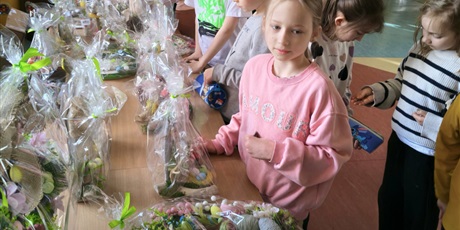 Powiększ grafikę: Grupa dzieci na korytarzu szkolnym podczas kiermaszu wielkanocnego. Dzieci oglądają i kupują ozdoby wielkanocne. Na stole rozłożone są dekoracje.