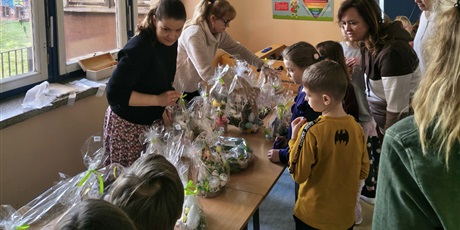 Powiększ grafikę: Grupa dzieci na korytarzu szkolnym podczas kiermaszu wielkanocnego. Dzieci oglądają i kupują ozdoby wielkanocne. Na stole rozłożone są dekoracje. Dwie nauczycielki sprzedają dekoracje.