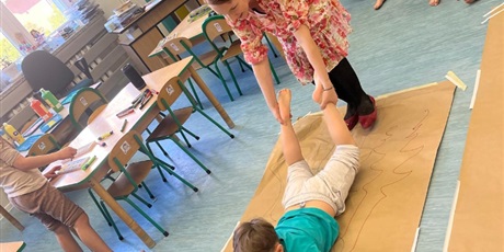 Powiększ grafikę: Chłopiec maluje obraz na szarym arkuszu papieru. Jest trzymany za nogi  przez nauczycielkę i przesuwany po papierze.