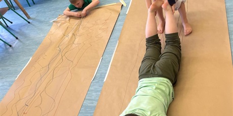 Powiększ grafikę: Chłopiec maluje obraz na szarym arkuszu papieru. Jest trzymany za nogi  przez koleżankę i przesuwany po papierze.