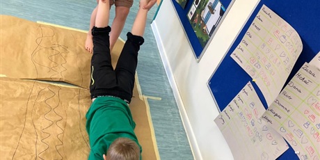 Powiększ grafikę: Chłopiec maluje obraz na szarym arkuszu papieru. Jest trzymany za nogi  przez kolegę i przesuwany po papierze.