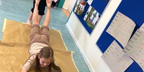 Powiększ grafikę: Dziewczynka maluje obraz na szarym arkuszu papieru. Jest trzymana za nogi  przez kolegę i przesuwana po papierze.