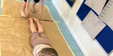 Powiększ grafikę: Dziewczynka maluje obraz na szarym arkuszu papieru. Jest trzymana za nogi  przez kolegę i przesuwana po papierze.