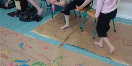 Powiększ grafikę: Dzieci siedzą na krzesełkach i czyszczą stopy z farby. Przed chwilą malowali obraz stopami umoczonymi w kolorowych farbkach.