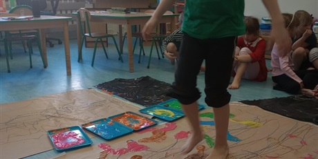 Powiększ grafikę: Chłopiec chodzi po arkuszu szarego papieru bosymi stopami. Stopy ma umoczone w farbie. Tworzy obraz.