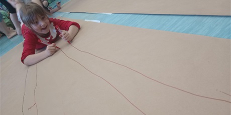 Powiększ grafikę: Dziewczynka maluje obraz na szarym papierze mazakami. Jest bardzo szczęśliwa i uśmiechnięta.