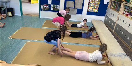 Powiększ grafikę: Dzieci leżą na podłodze na przuchu. Pod nii jest szary papier. W ręku maja mazaki. Inne dzieci unoszą dzieci za nogi. Będą je przesuwać po papierze. Powstanie obraz.