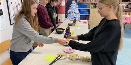 Powiększ grafikę: Zdjęcie ukazuje po lewej stronie uczniów, którzy prezentują swoje potrawy. Pierwsza dziewczyna podaje na talerzyku pieroga koleżance