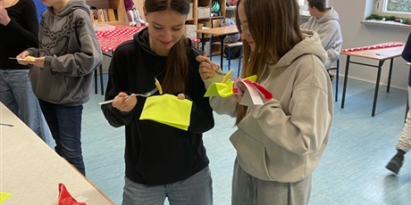 Powiększ grafikę: Dwie uczennice jedzą pierogi. W tle ukazani są inni uczniowie zastanawiający się nad oceną skosztowanych przed chwilą potraw