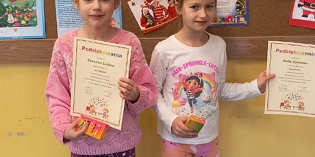 Powiększ grafikę: Uczennice stoją w klasie, w tle tablica. Prezentują dyplomy.