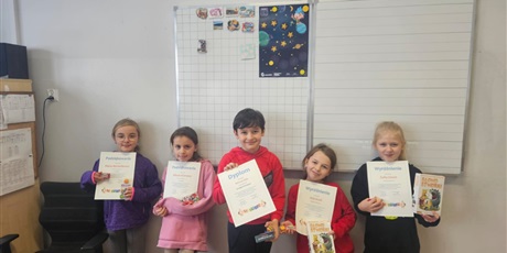 Powiększ grafikę: Uczniowie stoją obok siebie, w tle tablica. Prezentują dyplomy i nagrody. Uśmiechają się.