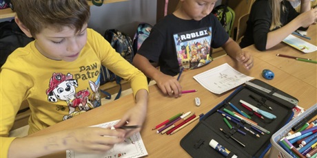 Powiększ grafikę: Dwóch chłopców i dziewczynka siedzą przy stole w świetlicy. W rękach trzymają kredki i wykonują projekty znaczków pocztowych. Na stole znajdują się pojemniki z kradkami i piórniki.