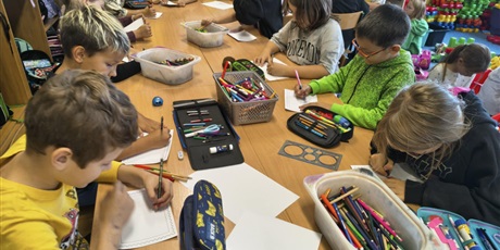 Powiększ grafikę: Grupa dzieci siedzi przy stole w świetlicy. W rękach trzymają kredki i wykonują projekty znaczków pocztowych. Na stole znajdują się pojemniki z kradkami i piórniki.