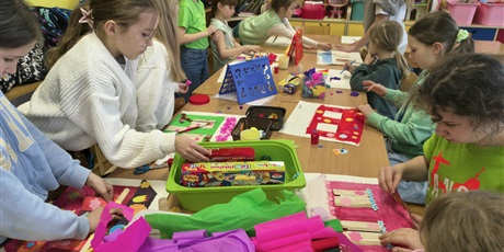 Powiększ grafikę: Grupa dzieci siedzi lub stoi przy stole i wykonuje swoje prace konkursowe. Na stole leżą kolorowe pojemniki, bibuła, kredki, farby.