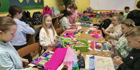 Powiększ grafikę: Grupa dzieci siedzi przy stole i wykonuje swoje prace konkursowe. Na stole leżą kolorowe pojemniki, bibuła, kredki, farby.