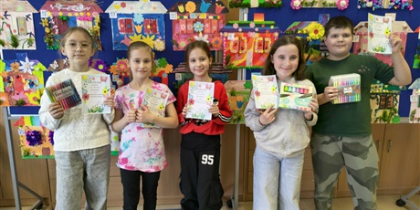 Powiększ grafikę:  Grupa dzieci pozuje do zdjęcia. W rękach trzymają dyplomy i nagrody za udział w konkursie. Za nimi znajduje się tablica, na której wiszą kolorowe prace plastyczne przedstawiające domy pani wiosny.