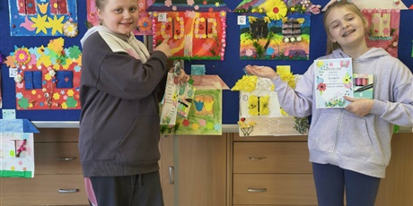 Powiększ grafikę: Dwie dziewczynki pozują do zdjęcia wskazując swoje prace konkursowe. W rękach trzymają dyplomy i nagrody. Za nimi znajduje się tablica, na której wiszą kolorowe prace plastyczne przedstawiające domy p