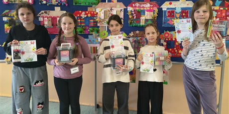 Powiększ grafikę: Grupa dzieci pozuje do zdjęcia. W rękach trzymają dyplomy i nagrody za udział w konkursie. Za nimi znajduje się tablica, na której wiszą kolorowe prace plastyczne przedstawiające domy pani wiosny.