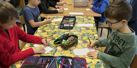 Powiększ grafikę: Grupa chłopców siedzi przy stole w świetlicy i maluje pracę konkursową pt. "Kwiatek dla każdej koleżanki". Na stole stoją kartki i pojemniki z mazakami i kredkami.