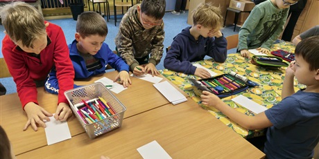 Powiększ grafikę: Grupa chłopców siedzi przy stole w świetlicy i maluje pracę konkursową pt. "Kwiatek dla każdej koleżanki". Na stole stoją kartki i pojemniki z mazakami i kredkami.