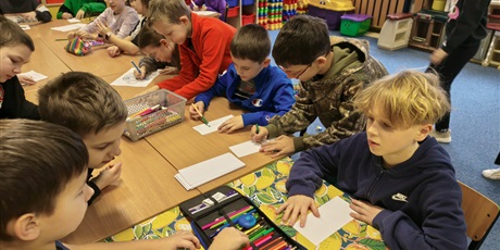 Powiększ grafikę: Grupa chłopców siedzi przy stole w świetlicy i maluje pracę konkursową pt. "Kwiatek dla każdej koleżanki". Na stole stoją kartki i pojemniki z mazakami i kredkami.