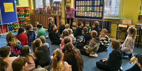 Powiększ grafikę: Grupa dzieci siedzi na dywanie w świetlicy. Dzieci patrzą na nauczycielkę, która pokazuje prace konkursowe wiszące na granatowej tablicy.