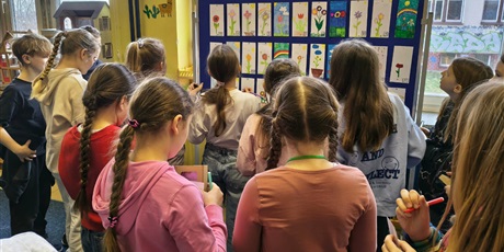 Powiększ grafikę: Grupa dziewczynek przygląda się pracom konkursowym wiszącym na granatowej tablicy. W dłoni trzymają kartki do głosowania i ołówki. 