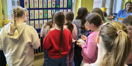 Powiększ grafikę: Grupa dziewczynek przygląda się pracom konkursowym wiszącym na granatowej tablicy. W dłoni trzymają kartki do głosowania i ołówki.