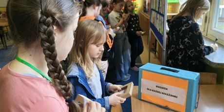 Powiększ grafikę: Grupa dziewczynek przygląda się pracom konkursowym wiszącym na granatowej tablicy. W dłoni trzymają kartki do głosowania i ołówki. Dwie dziewczynki wrzucają swoje losy do niebieskiej skrzynki.