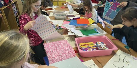 Powiększ grafikę: Grupa dzieci siedzi przy stole w świetlicy i wykleja postać kota. Na stole leżą pojemniki z kolorowymi kartkami, piankami, filcem, kredkami itp.