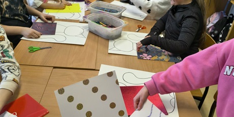 Powiększ grafikę: Grupa dzieci siedzi przy stole w świetlicy i wykleja postać kota. Na stole leżą pojemniki z kolorowymi kartkami, piankami, filcem, kredkami itp.