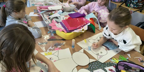 Powiększ grafikę: Grupa dzieci siedzi przy stole w świetlicy i wykleja postać kota. Na stole leżą pojemniki z kolorowymi kartkami, piankami, filcem, kredkami itp.